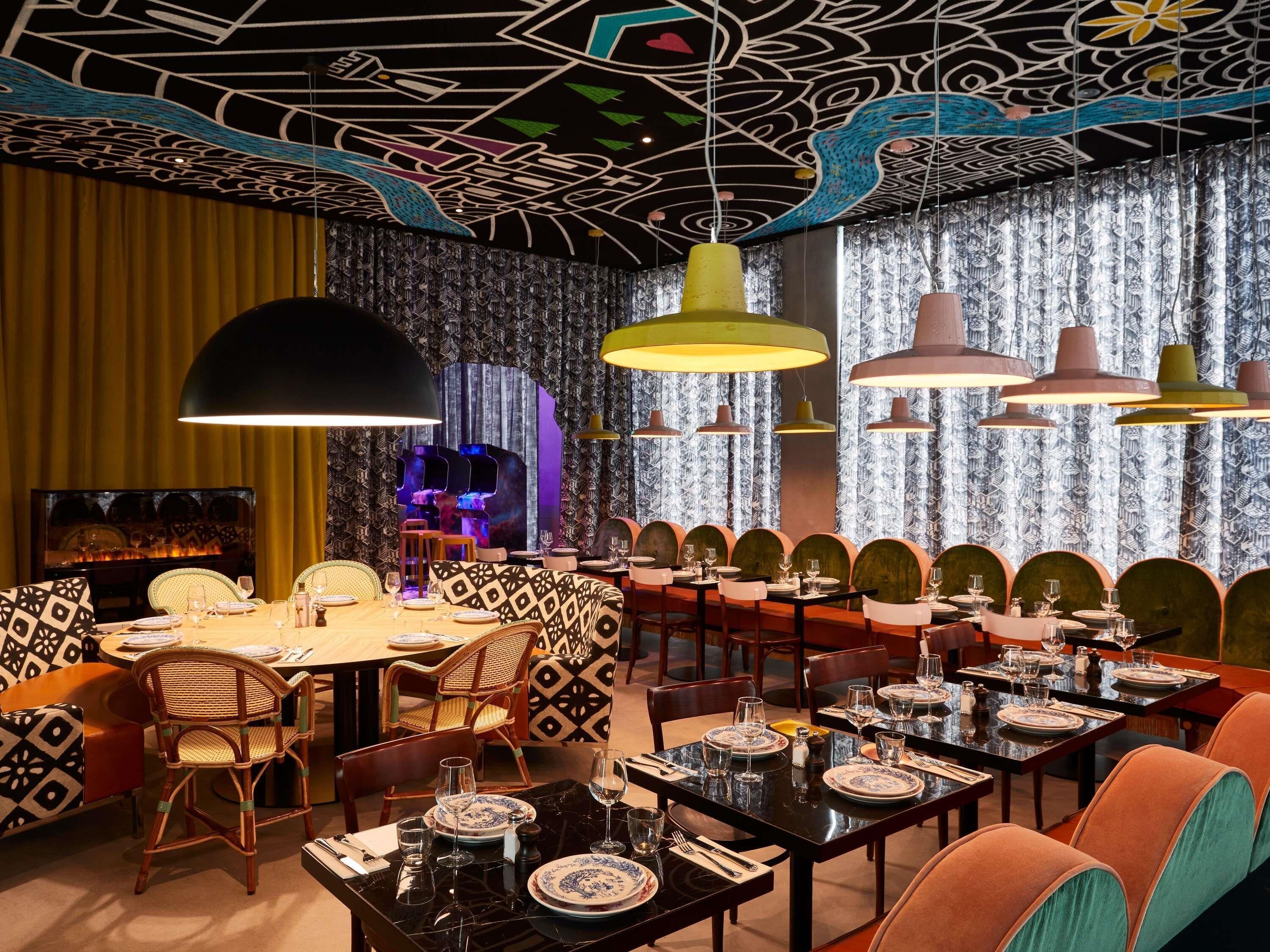 Un restaurante con una decoración moderna, que destaca por un papel tapiz estampado, arte único en el techo y diversas opciones de asientos.