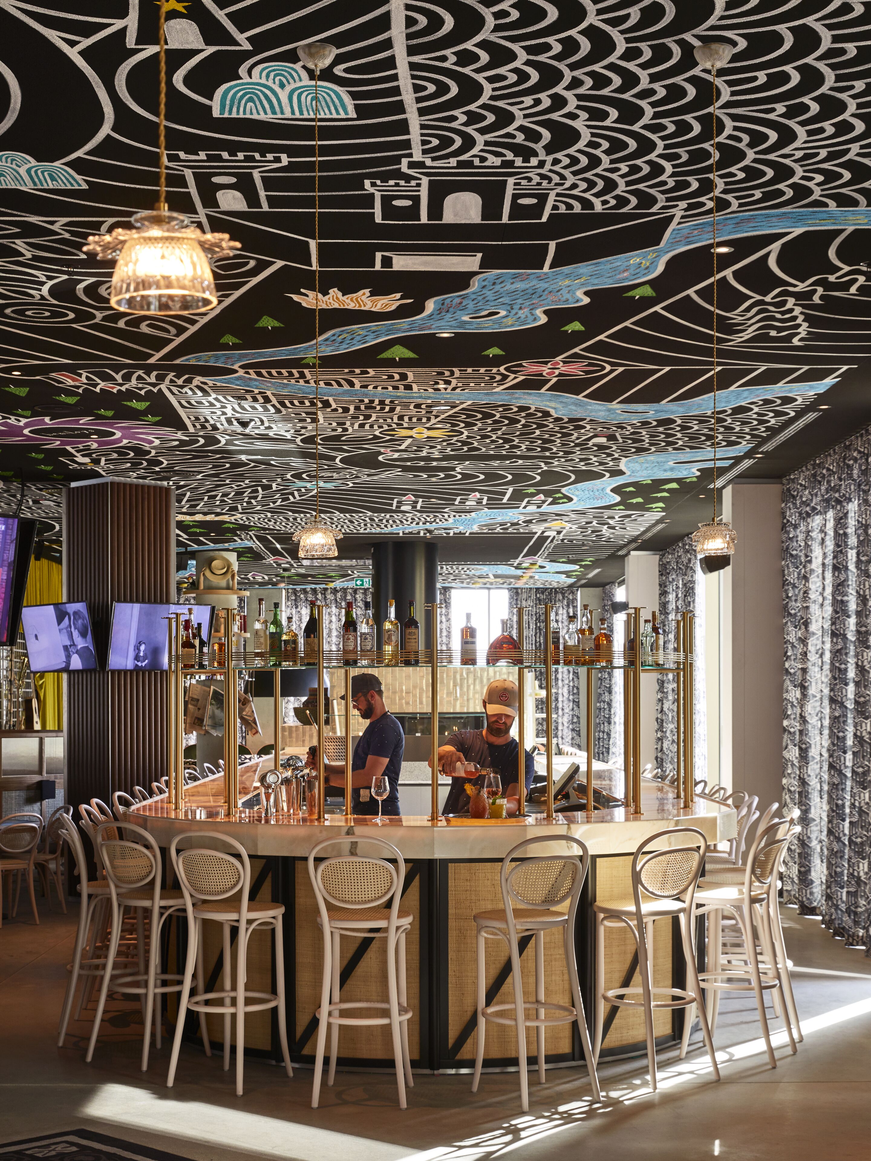 Een bar met een uniek plafondontwerp, barstoelen en een counter waar een bartender drankjes aan het bereiden is.