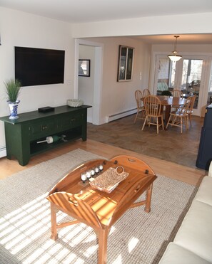 A living room with a wooden coffee table, a green entertainment unit, a flat-screen TV, and a dining area with wooden chairs and a table.
