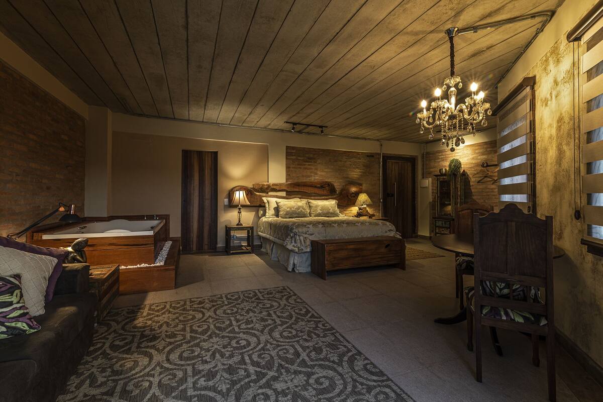 Ein gemütliches Schlafzimmer mit Holzbalkendecke, einem großen Bett, einer Holzkommode, einem Sessel und einer Deckenleuchte.