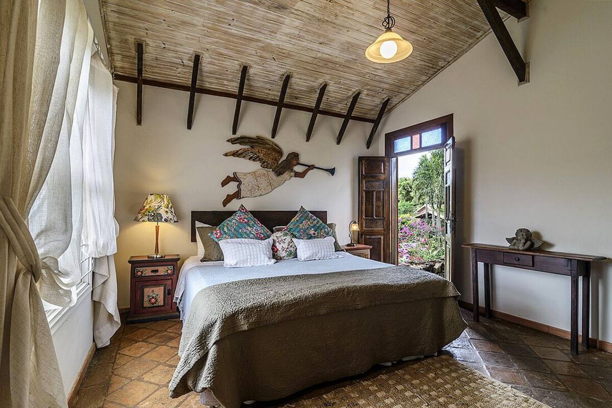 Ein Schlafzimmer mit einem großen Bett, Holzbalken an der Decke, einem Nachttisch mit Lampe und einem Fenster mit Blick auf Grünflächen.