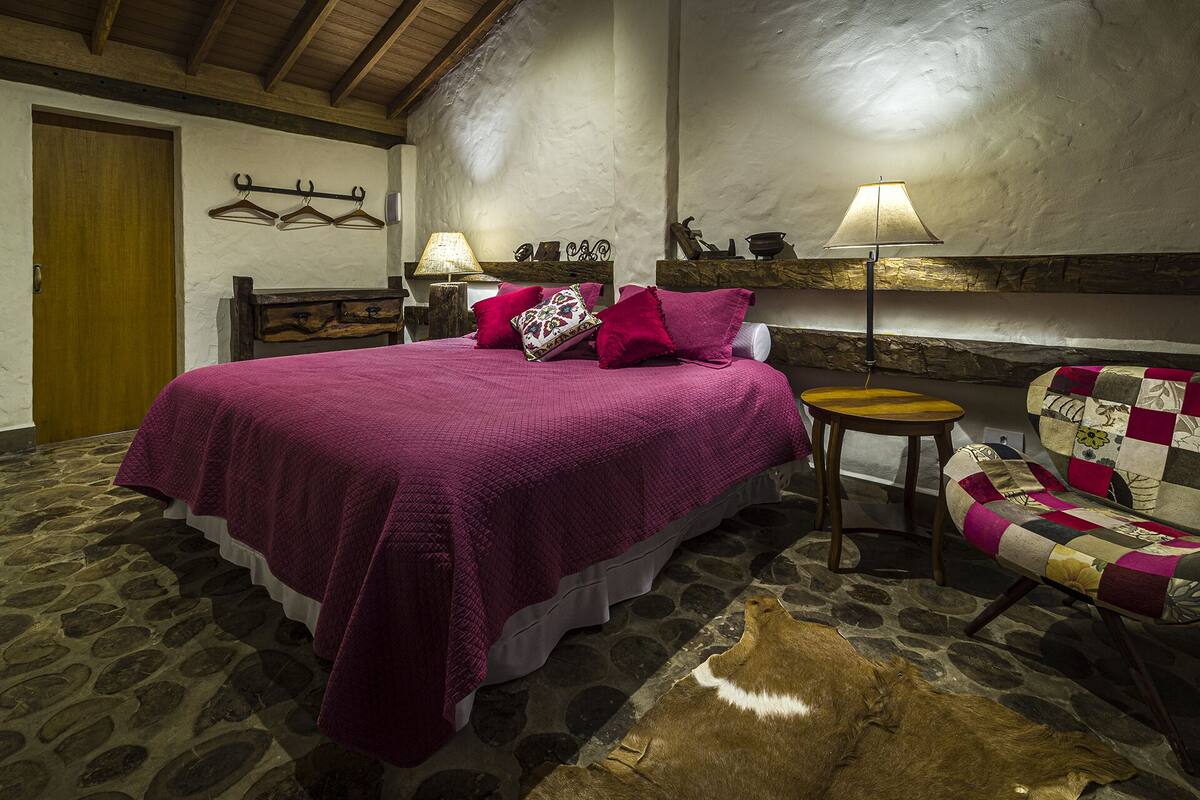 Ein Schlafzimmer mit einem großen Bett, einem Holzkopfende, Steinböden, einer Holztür, einem Holzstuhl, einer Lampe und einem an der Wand befestigten Kleiderhaken.
