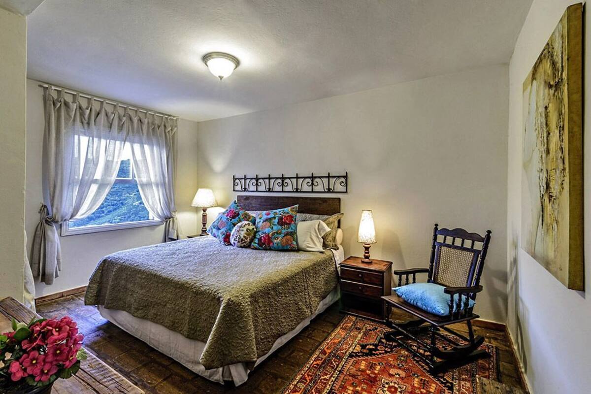 Ein Schlafzimmer mit einem großen Bett, einem Nachttisch mit Lampe, einem Schaukelstuhl, einem Fenster mit Vorhängen und einem Wandbild.