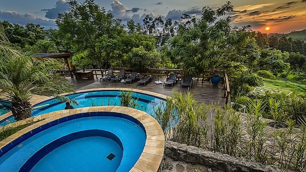 A swimming pool with a wooden deck and lounge chairs, surrounded by lush greenery and a sunset view.
