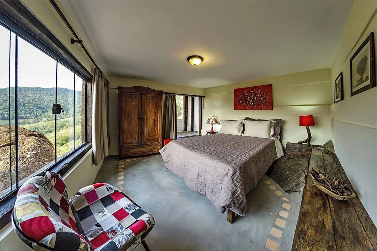 Ein Schlafzimmer mit einem großen Bett, einem Holzschrank, einem Sessel, einem Tisch mit Lampe und Blick ins Freie durch ein Fenster.