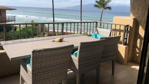 Un balcón con mesa de madera y sillas de mimbre, con vistas al océano, la costa y palmeras.
