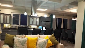 Un soggiorno con un divano color beige, numerosi cuscini, una zona pranzo con sedie e una cucina sullo sfondo.