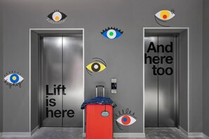 Un ascenseur dans lequel se trouve une valise rouge, et un écran numérique affiche le chiffre « 50 ».