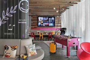 A modern hotel lobby with a foosball table, a sofa with cushions, a television displaying multiple screens, and a wooden ceiling. - Moxy Bucharest Old Town (Bucharest)