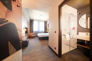 Habitación de hotel con cama, escritorio, silla, ducha y baño con lavamanos y espejo.