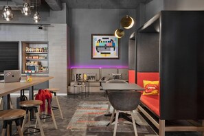 A modern living space with a sofa, a dining table, and bar stools. There is a laptop on the table, and a shelf with various items. - Moxy Bucharest Old Town (Bucharest)