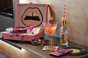 Un tavolo con una valigia rosa, un bicchiere da martini con una cannuccia con la scritta 'Cheers', una bottiglia d'acqua, un paio di occhiali e una cover glitterata per telefono su un vinile.