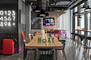 A modern interior with a long wooden table, chairs, a red suitcase, and a digital display with numbers. - Moxy Bucharest Old Town (Bucharest)