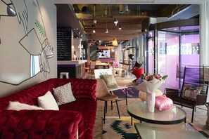 A modern interior with a red velvet sofa, a glass coffee table, and a bar area in the background. - Moxy Bucharest Old Town (Bucharest)
