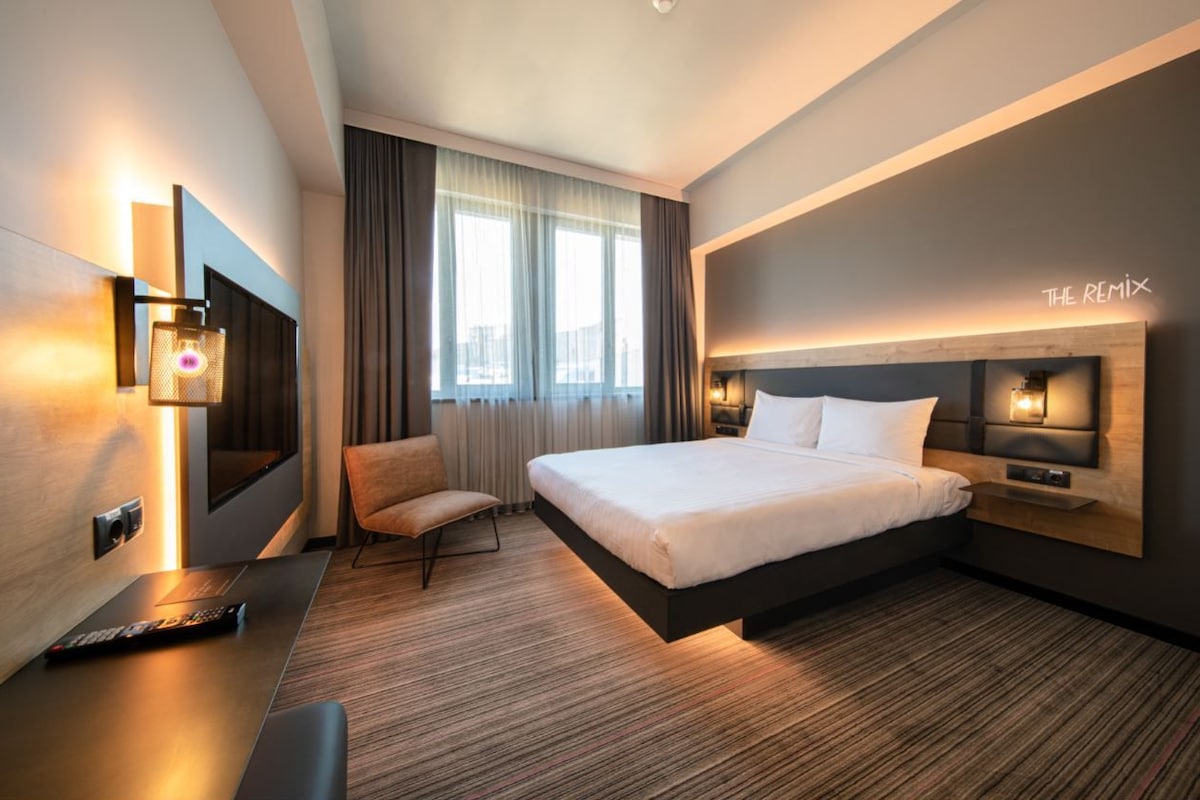 A modern hotel room with a large bed, a desk with a remote control, a chair, and a TV mounted on the wall.