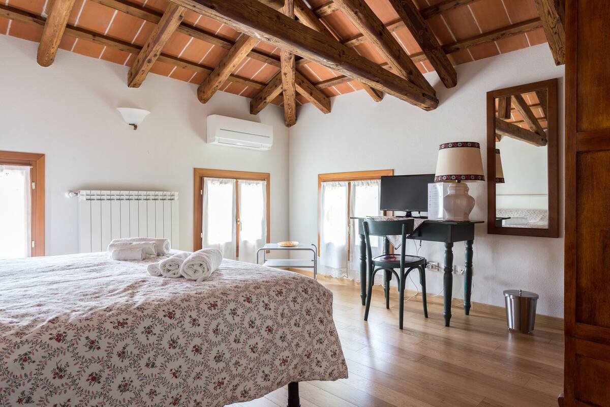 Ein Schlafzimmer mit Holzbalkendecke, einem Bett mit floralem Überwurf, einem Schreibtisch mit Lampe und einem Spiegel an der Wand.