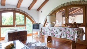 Une pièce rustique avec des poutres en bois, un téléviseur, une table recouverte d’une nappe à motifs floraux et une grande fenêtre donnant sur la forêt.
