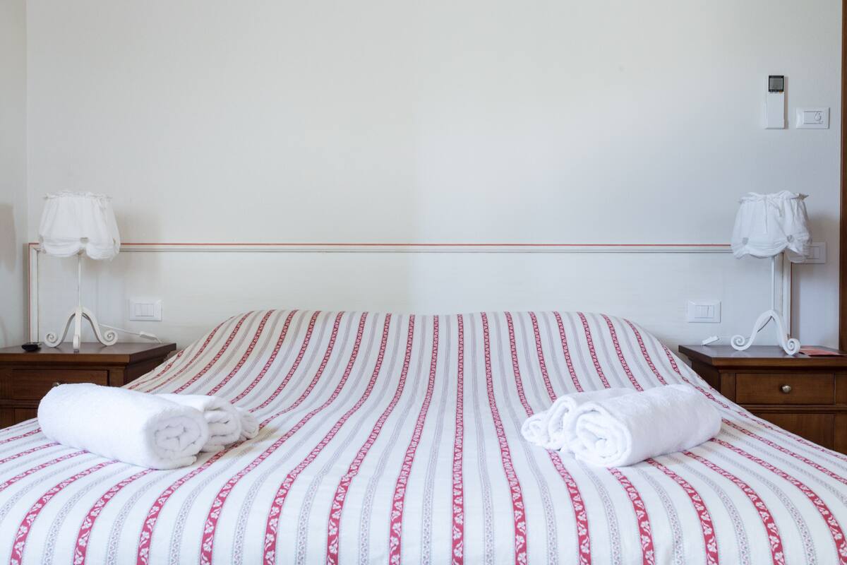 Ein ordentlich gemachtes Bett mit gestreifter Bettdecke, zwei Nachttische mit Lampen und einem an der Wand befestigten Lichtschalter.