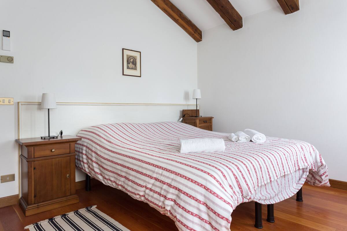 Ein Schlafzimmer mit einem Bett, das mit einer gestreiften Bettdecke bezogen ist, einem Nachttisch aus Holz mit einer Lampe, einem kleinen Schrank und einem gerahmten Bild an der Wand.
