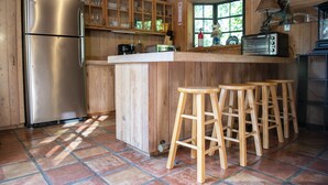 A wooden kitchen with a stainless steel refrigerator, wooden stools, and a window with a view of greenery. - Quaint Pool House in Tropical Setting (Boynton Beach)