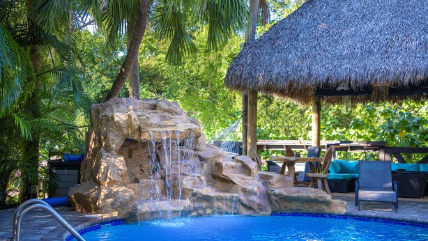 A pool with a rock waterfall, surrounded by lush greenery and a thatched-roof structure. - Quaint Pool House in Tropical Setting (Boynton Beach)
