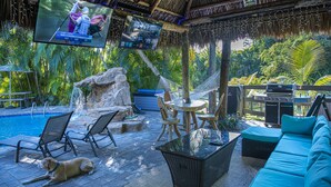 A poolside area with lounge chairs, a sofa, a TV screen, a grill, and a dog. - Quaint Pool House in Tropical Setting (Boynton Beach)