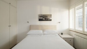 Een slaapkamer met een bed, nachtkastje, kledingkast en een raam met jaloezieën.