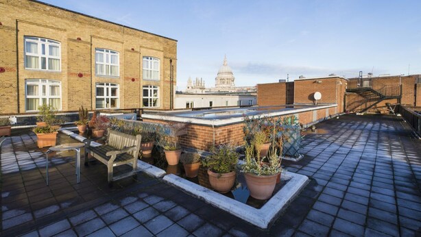 Een dakterras met plantenbakken, een houten bank en uitzicht op een historisch gebouw.