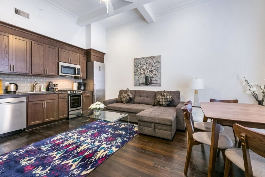 Una cocina moderna con armarios de madera, un microondas y una estufa. Hay un sofá gris con cojines estampados y una mesa de comedor con sillas. Una alfombra colorida cubre el suelo.
