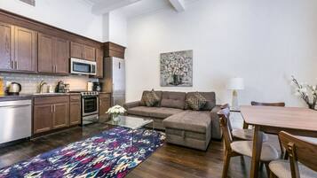 Una cocina moderna con armarios de madera, un microondas y una estufa. Hay un sofá gris con cojines estampados y una mesa de comedor con sillas. Una alfombra colorida cubre el suelo.