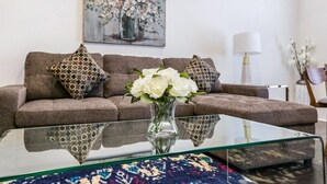 A living room with a brown sofa, patterned pillows, a glass coffee table, and a vase with white flowers.