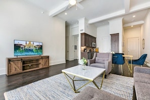 A modern living room with a flat-screen TV, a coffee table, and a sofa. There is a kitchen area in the background with cabinets and a microwave.