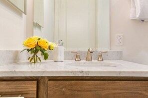 Un baño con lavamanos de mármol, dos grifos dorados, dispensador de jabón blanco y un jarrón con rosas amarillas.