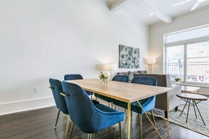 A modern living room with a wooden dining table, blue velvet chairs, a grey sofa, and a large window providing natural light.
