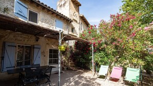 A rustic stone building with a patio area featuring a table and chairs, a hanging plant, and a tree with pink flowers. - Gîte du Pigeonnier, 3 Épis, Mas Médaille (arles)