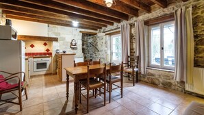 A rustic kitchen with stone walls, wooden beams, a dining area with a wooden table and chairs, and a view of the outdoors through a window. - Gîte du Pigeonnier, 3 Épis, Mas Médaille (arles)