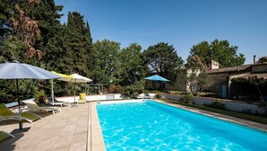 A swimming pool with clear blue water, surrounded by lounge chairs and umbrellas, set against a backdrop of trees and a building. - Gîte du Pigeonnier, 3 Épis, Mas Médaille (arles)