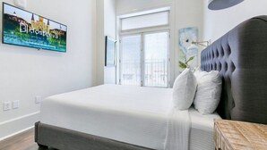 A modern hotel room with a large bed, a television displaying the hotel's name, a window with blinds, and a decorative lamp. - Stunning Canal St Views • Steps to Bourbon (New Orleans)