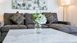 A living room with a grey sofa, patterned cushions, a vase with flowers, and a floor lamp. - Stunning Canal St Views • Steps to Bourbon (New Orleans)