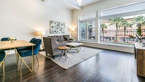 A modern living room with a dark wood floor, a sofa, a coffee table, and a dining table with chairs. There is a large window with a view of palm trees and a fence. - Stunning Canal St Views • Steps to Bourbon (New Orleans)