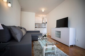 Una sala de estar moderna con un sofá gris, una mesa de centro de vidrio con una botella y vasos, un televisor de pantalla plana sobre un mueble blanco, y una cocina al fondo.