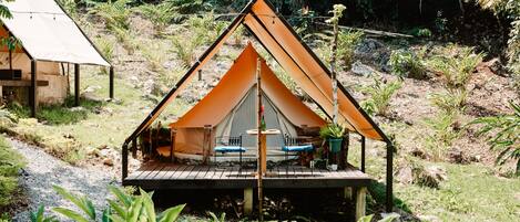 A small, triangular tent with a wooden deck and two chairs, surrounded by greenery.