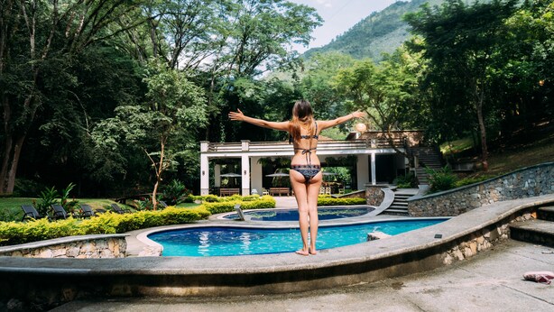 Una mujer de pie junto a una piscina, con vista a un edificio y árboles al fondo.