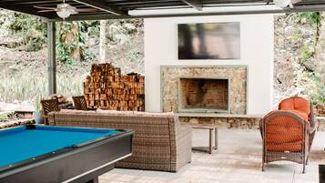 An outdoor patio with a pool table, wicker furniture, a stone fireplace, and a TV mounted on the wall.