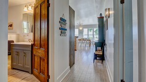 Un pasillo con una puerta de madera, un lavamanos y un espejo. Se ven sillas y una mesa al fondo.