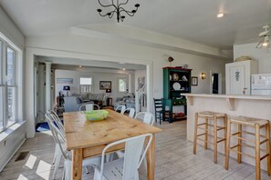 A spacious dining area with a wooden table and white chairs, a kitchen with a refrigerator and cabinets, and a view of the living room through an open doorway.