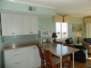 A kitchen with white cabinets, a microwave, and a dining area with a table and chairs. -  Wake up to the Surises and soothing sound of waves in this ocean front condo. (North Myrtle Beach)