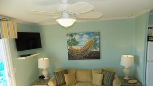 Uma sala de estar com um sofá bege, duas mesinhas de cabeceira com luminárias, um ventilador de teto, uma televisão na parede e uma pintura de rede.