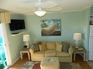 A living room with a beige sofa, two side tables with lamps, a ceiling fan, a mounted TV, and a hammock painting on the wall. -  Wake up to the Surises and soothing sound of waves in this ocean front condo. (North Myrtle Beach)