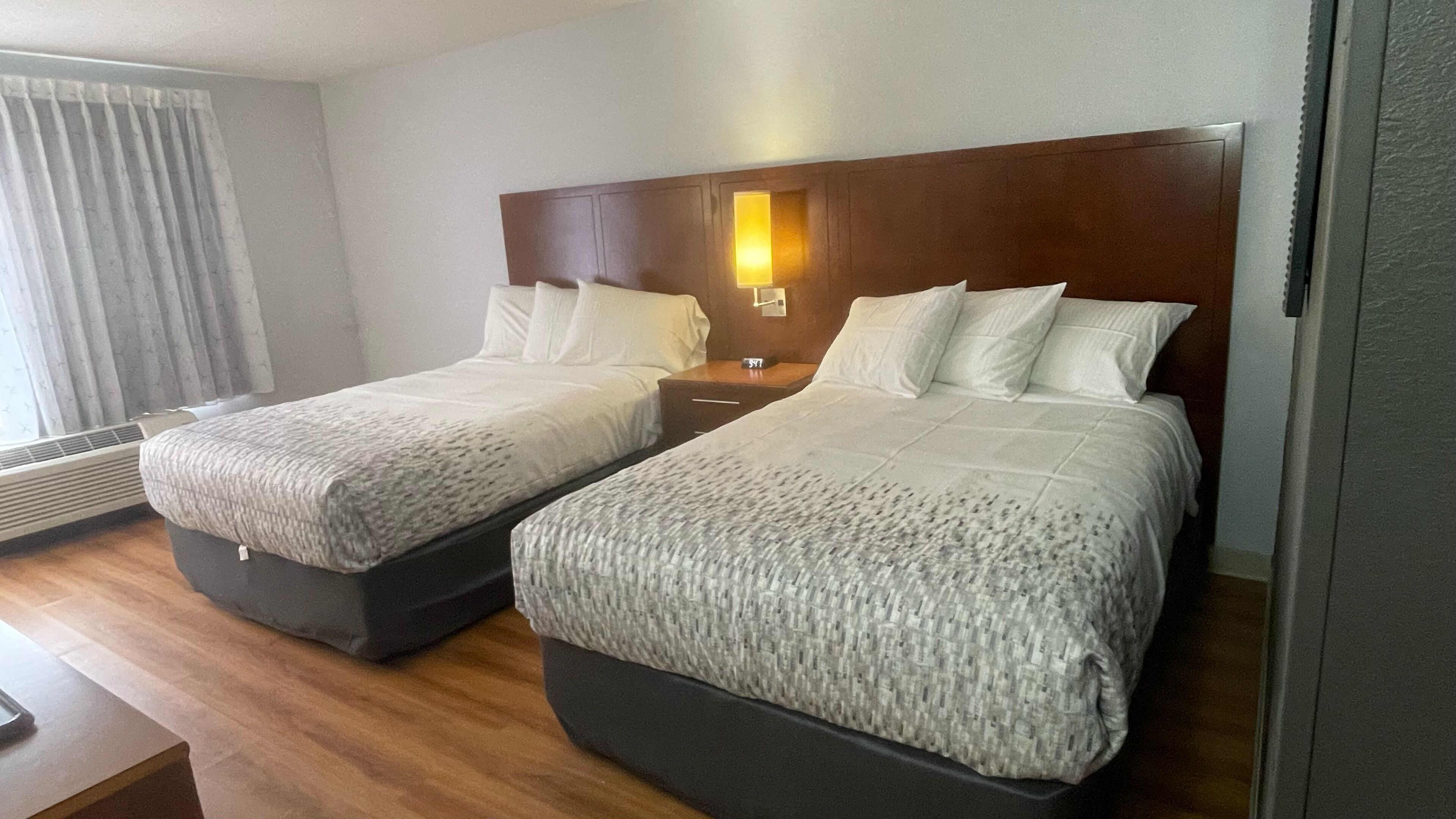 Une chambre d’hôtel avec deux lits, une tête de lit en bois, une table de chevet avec une lampe et une fenêtre avec des rideaux.